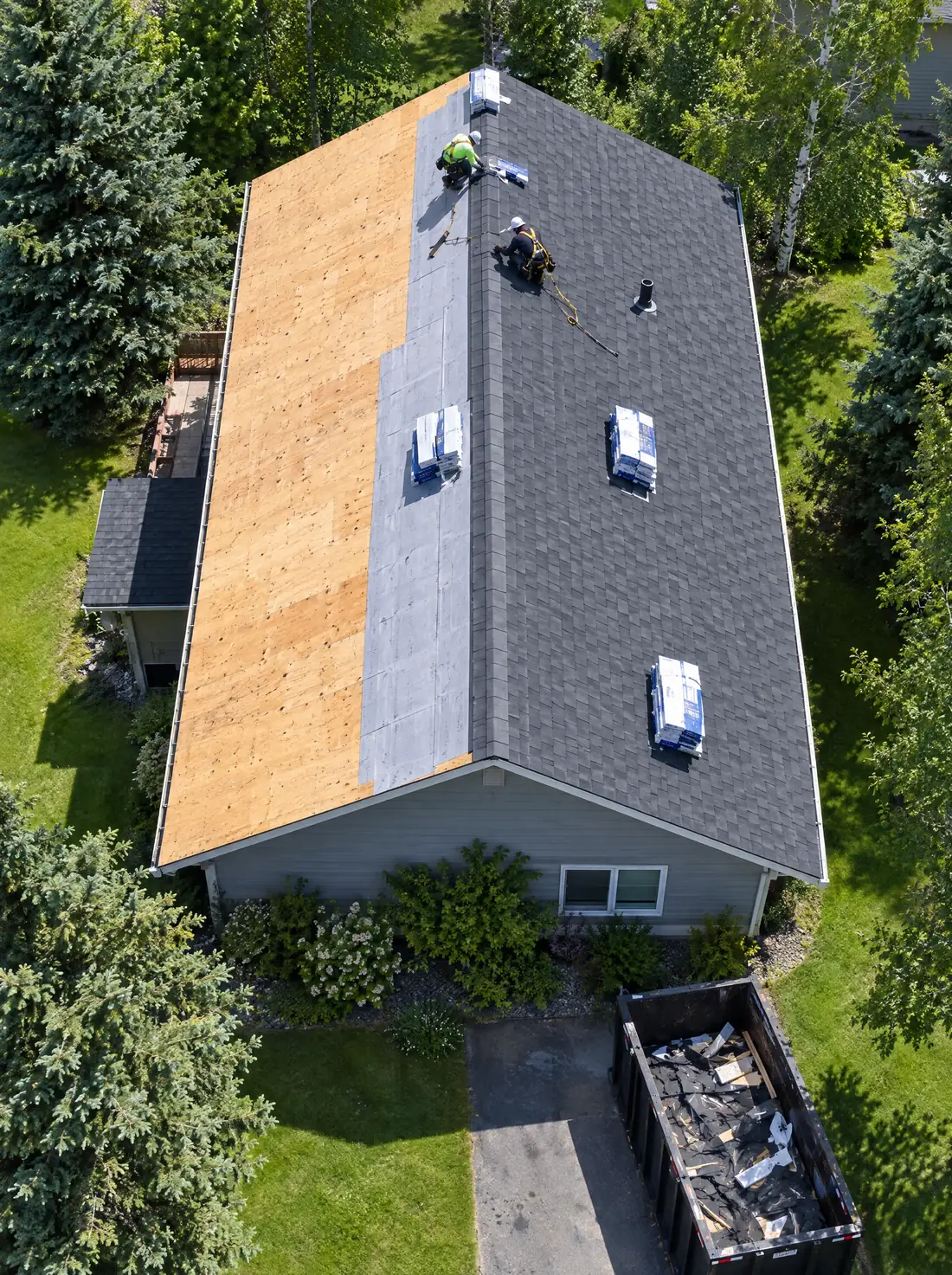 A roof seen from above in July, half stripped to plywood and half in new shingles, two roofers near the ridge